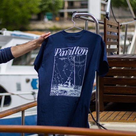 A hand is holding a navy blue T-shirt with a design featuring a boat and navigation lines, along with the word 'Pavillon.' The background includes a boat deck with wooden furniture and a partially visible white and blue boat.