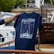 A hand is holding a navy blue T-shirt with a design featuring a boat and navigation lines, along with the word 'Pavillon.' The background includes a boat deck with wooden furniture and a partially visible white and blue boat.