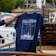 A hand is holding a navy blue T-shirt with a design featuring a boat and navigation lines, along with the word 'Pavillon.' The background includes a boat deck with wooden furniture and a partially visible white and blue boat.