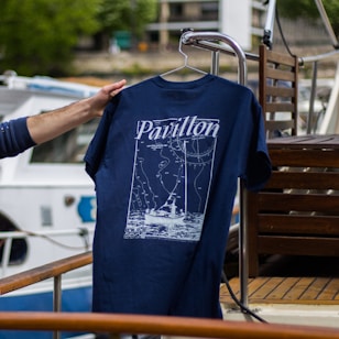 A hand is holding a navy blue T-shirt with a design featuring a boat and navigation lines, along with the word 'Pavillon.' The background includes a boat deck with wooden furniture and a partially visible white and blue boat.