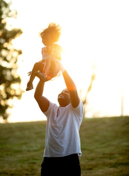 A man joyfully lifts a child into the air against a bright, sunlit background. Both are outdoors in a park-like setting, and the man's expression appears happy. The child is wearing a patterned dress and shoes.