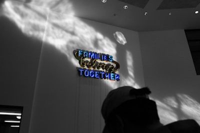 Neon signage with the words 'Families Belong Together' is mounted on a wall, illuminated in bright colors. Shadows cast dramatic patterns on the wall, and the scene includes a silhouette of a person wearing a cap in the foreground.