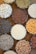 Circular bowls filled with various dried grains and legumes such as lentils, rice, beans, corn, sunflower seeds, chickpeas, and others are arranged on a flat surface. The bowls are placed closely together, creating a pattern of different colors and textures.