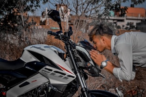 A person is shown leaning towards a motorcycle, gently touching and appearing to kiss the front part of the vehicle. The motorcycle is a sporty model with a sleek design, predominantly in white and black, and features the brand name on the side. The setting seems to be outdoors with dry vegetation and a blurred urban background.