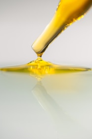 A dropper releases a viscous yellow liquid onto a smooth, reflective surface. The liquid appears to be oil-like and spreads gently, creating ripples and reflections on the surface below.