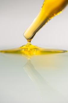 A dropper releases a viscous yellow liquid onto a smooth, reflective surface. The liquid appears to be oil-like and spreads gently, creating ripples and reflections on the surface below.