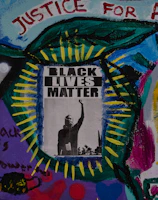 A vibrant mural featuring a central poster with the words 'Black Lives Matter' and an image of a raised fist. Surrounding the poster are colorful abstract patterns and words like 'Justice for All,' 'Black is Power,' and 'Shine,' painted with various colors including blue, green, red, and purple. The artwork conveys a message of empowerment and social justice.