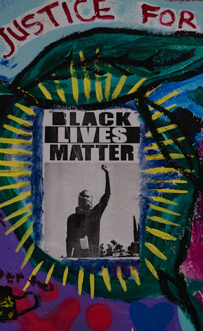 A vibrant mural featuring a central poster with the words 'Black Lives Matter' and an image of a raised fist. Surrounding the poster are colorful abstract patterns and words like 'Justice for All,' 'Black is Power,' and 'Shine,' painted with various colors including blue, green, red, and purple. The artwork conveys a message of empowerment and social justice.