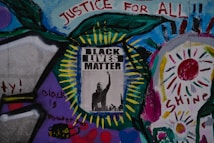 A vibrant mural featuring a central poster with the words 'Black Lives Matter' and an image of a raised fist. Surrounding the poster are colorful abstract patterns and words like 'Justice for All,' 'Black is Power,' and 'Shine,' painted with various colors including blue, green, red, and purple. The artwork conveys a message of empowerment and social justice.