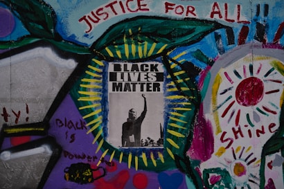 A vibrant mural featuring a central poster with the words 'Black Lives Matter' and an image of a raised fist. Surrounding the poster are colorful abstract patterns and words like 'Justice for All,' 'Black is Power,' and 'Shine,' painted with various colors including blue, green, red, and purple. The artwork conveys a message of empowerment and social justice.