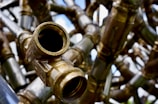 A detailed and close-up view of interconnected brass pipes with a large open valve facing towards the viewer. The shiny metallic surfaces reflect light, creating a complex network of lines and curves. The background is slightly out of focus, adding depth to the intricate pattern of pipes.