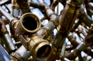 A detailed and close-up view of interconnected brass pipes with a large open valve facing towards the viewer. The shiny metallic surfaces reflect light, creating a complex network of lines and curves. The background is slightly out of focus, adding depth to the intricate pattern of pipes.