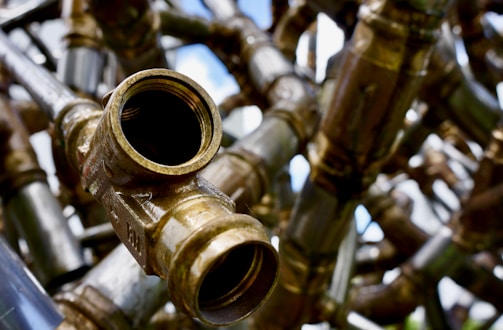 A detailed and close-up view of interconnected brass pipes with a large open valve facing towards the viewer. The shiny metallic surfaces reflect light, creating a complex network of lines and curves. The background is slightly out of focus, adding depth to the intricate pattern of pipes.
