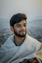 A person with a short beard and a gentle smile is wrapped in a light-colored blanket with a blurred cityscape in the background. The setting appears to be outdoors with a serene and peaceful atmosphere.