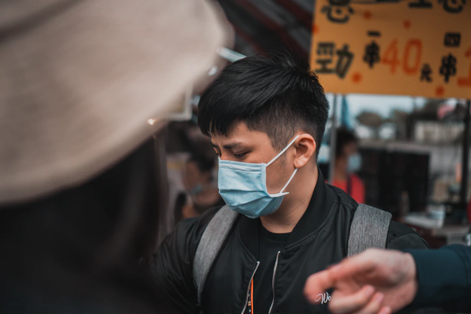 The image shows a man wearing a face mask, possibly due to an ongoing pandemic or health concern. He is standing outdoors and appears to be engaged in conversation with another person who is also wearing a face mask. The background includes a sign that reads "40", which could potentially indicate the price of something nearby.