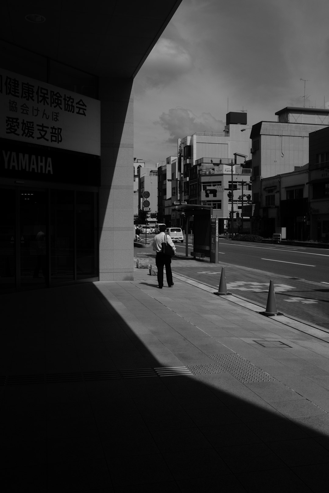 grayscale photo of man in black jacket and pants walking on sidewalk