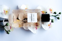 A Chanel perfume bottle lying horizontally against a backdrop of white orchids with pink and yellow centers. The transparent bottle reveals a light amber liquid inside, adorned with delicate bubbles and petals. The label reads 'Paris - Venise Chanel'. The composition is elegant and luxurious.