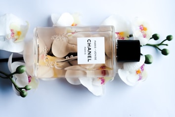 A Chanel perfume bottle lying horizontally against a backdrop of white orchids with pink and yellow centers. The transparent bottle reveals a light amber liquid inside, adorned with delicate bubbles and petals. The label reads 'Paris - Venise Chanel'. The composition is elegant and luxurious.