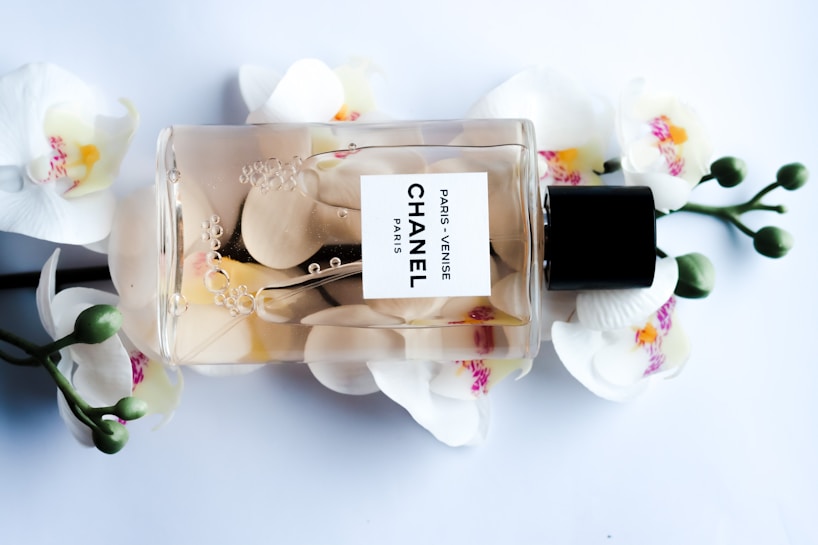 A Chanel perfume bottle lying horizontally against a backdrop of white orchids with pink and yellow centers. The transparent bottle reveals a light amber liquid inside, adorned with delicate bubbles and petals. The label reads 'Paris - Venise Chanel'. The composition is elegant and luxurious.