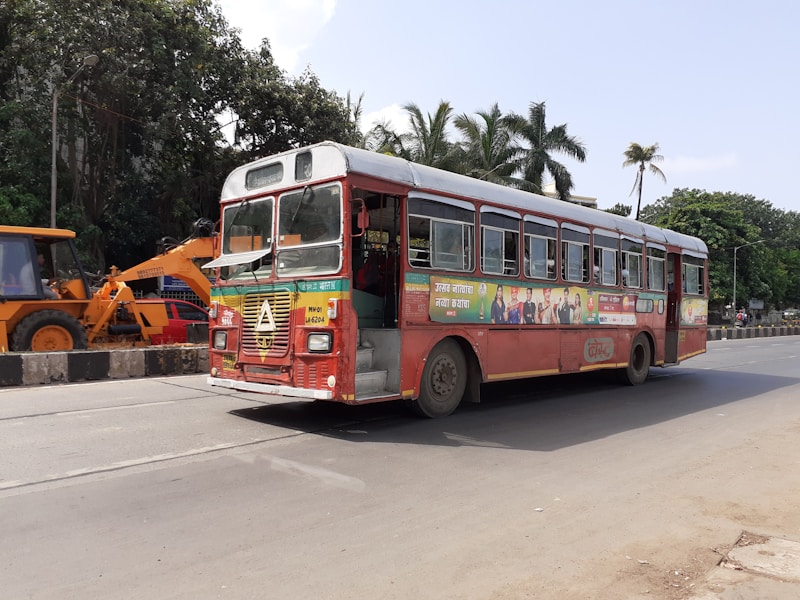 A red public bus is driving on a city street. The bus has a colorful advertisement along its side and is moving past a construction area where a yellow excavator is visible. Tall palm trees and lush green foliage line the roadside in the background. The sky is clear, suggesting a sunny day.