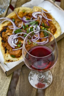 Close-up of a rustic wooden table with a glass of red wine and a pizza slice beside it, sunlight filtering through leaves.