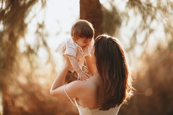 A woman with long brown hair is holding a laughing toddler up in the air. They are both outdoors, surrounded by soft-focus greenery, and the light creates a warm, golden atmosphere.