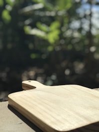 A wooden cutting board is placed on a surface with sunlight casting shadows. The background is blurred with green foliage indicating an outdoor setting.