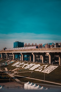A modern urban scene featuring a train traveling on an elevated track above a sprawling city park. Large white Chinese characters are displayed on a grassy hill. People are walking along the pathways, and a variety of tents and buildings are visible in the foreground and background. The sky is clear and turquoise, giving a sense of a bright day.
