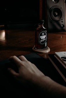 A dark, moody setting featuring a wooden desk with a cold brew coffee bottle labeled 'Morning Kicks' resting on a wooden coaster. A hand rests on a mouse pad, while a speaker with visible components sits in the background.