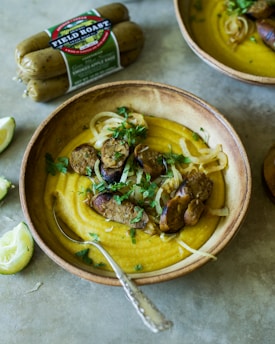 A rustic bowl filled with creamy yellow soup, topped with slices of browned sausage and garnished with herbs and onions. A metal spoon rests inside the bowl, and a package of Field Roast sausages is visible in the background. A squeezed lemon wedge sits on the table beside the bowl.