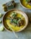 A rustic bowl filled with creamy yellow soup, topped with slices of browned sausage and garnished with herbs and onions. A metal spoon rests inside the bowl, and a package of Field Roast sausages is visible in the background. A squeezed lemon wedge sits on the table beside the bowl.