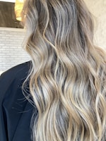 Wavy, long hair with blonde highlights cascades down against a background of light-colored brick wall and part of a black cape is visible.