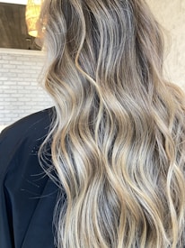 Wavy, long hair with blonde highlights cascades down against a background of light-colored brick wall and part of a black cape is visible.