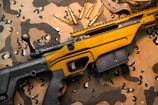 A black and gold rifle with the word 'Savage' engraved on the side rests on a camouflage fabric. Several gold bullets are arranged nearby, and protective goggles are partially visible in the upper right corner.