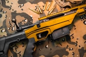 A black and gold rifle with the word 'Savage' engraved on the side rests on a camouflage fabric. Several gold bullets are arranged nearby, and protective goggles are partially visible in the upper right corner.