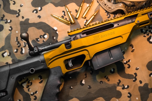 A black and gold rifle with the word 'Savage' engraved on the side rests on a camouflage fabric. Several gold bullets are arranged nearby, and protective goggles are partially visible in the upper right corner.