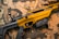 A black and gold rifle with the word 'Savage' engraved on the side rests on a camouflage fabric. Several gold bullets are arranged nearby, and protective goggles are partially visible in the upper right corner.