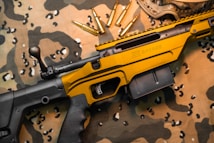 A black and gold rifle with the word 'Savage' engraved on the side rests on a camouflage fabric. Several gold bullets are arranged nearby, and protective goggles are partially visible in the upper right corner.