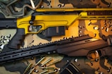 Two rifles are placed on a camouflage cloth surface. One rifle has a striking yellow finish, while the other is black. Scattered around them are several rifle cartridges and a pair of protective goggles.