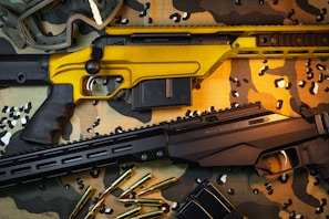 Two rifles are placed on a camouflage cloth surface. One rifle has a striking yellow finish, while the other is black. Scattered around them are several rifle cartridges and a pair of protective goggles.