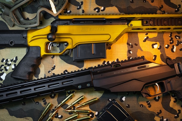Two rifles are placed on a camouflage cloth surface. One rifle has a striking yellow finish, while the other is black. Scattered around them are several rifle cartridges and a pair of protective goggles.