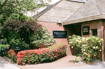 A brick building surrounded by lush greenery and vibrant flowers. The entrance is adorned with a sign reading 'Munro & Crawford, Barristers & Solicitors' and 'Notaries Public'. The address '5670 Yew Street' is displayed on a separate sign. There is a variety of foliage, including a large tree and shrubs with red and yellow flowers. A person is seated on a bench near the entrance, partially obscured by the plants.