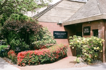 A brick building surrounded by lush greenery and vibrant flowers. The entrance is adorned with a sign reading 'Munro & Crawford, Barristers & Solicitors' and 'Notaries Public'. The address '5670 Yew Street' is displayed on a separate sign. There is a variety of foliage, including a large tree and shrubs with red and yellow flowers. A person is seated on a bench near the entrance, partially obscured by the plants.