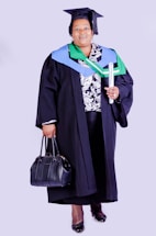 A person wearing academic regalia, including a graduation cap and gown, holding a rolled diploma in one hand and a black handbag in the other. The outfit is adorned with colorful stoles, indicating achievement or affiliation.