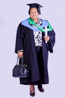 A person wearing academic regalia, including a graduation cap and gown, holding a rolled diploma in one hand and a black handbag in the other. The outfit is adorned with colorful stoles, indicating achievement or affiliation.