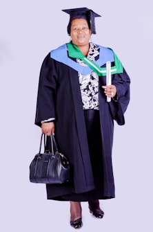 A person wearing academic regalia, including a graduation cap and gown, holding a rolled diploma in one hand and a black handbag in the other. The outfit is adorned with colorful stoles, indicating achievement or affiliation.