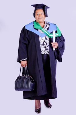 A person wearing academic regalia, including a graduation cap and gown, holding a rolled diploma in one hand and a black handbag in the other. The outfit is adorned with colorful stoles, indicating achievement or affiliation.