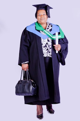 A person wearing academic regalia, including a graduation cap and gown, holding a rolled diploma in one hand and a black handbag in the other. The outfit is adorned with colorful stoles, indicating achievement or affiliation.