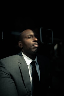 A man in a suit is partially illuminated by light in a dark setting. The focus is on his contemplative expression as he gazes upward. His attire includes a white shirt, dark tie, and a grey suit, reflecting a professional appearance.