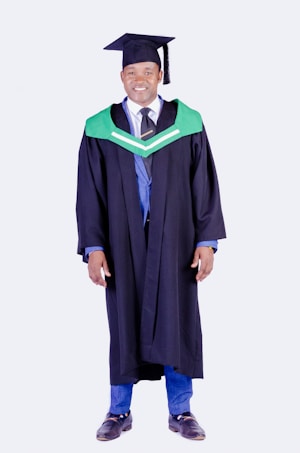 A person wearing a graduation gown and cap, featuring a green and black color scheme with a tassel. The attire includes a formal shirt, tie, and blue pants.